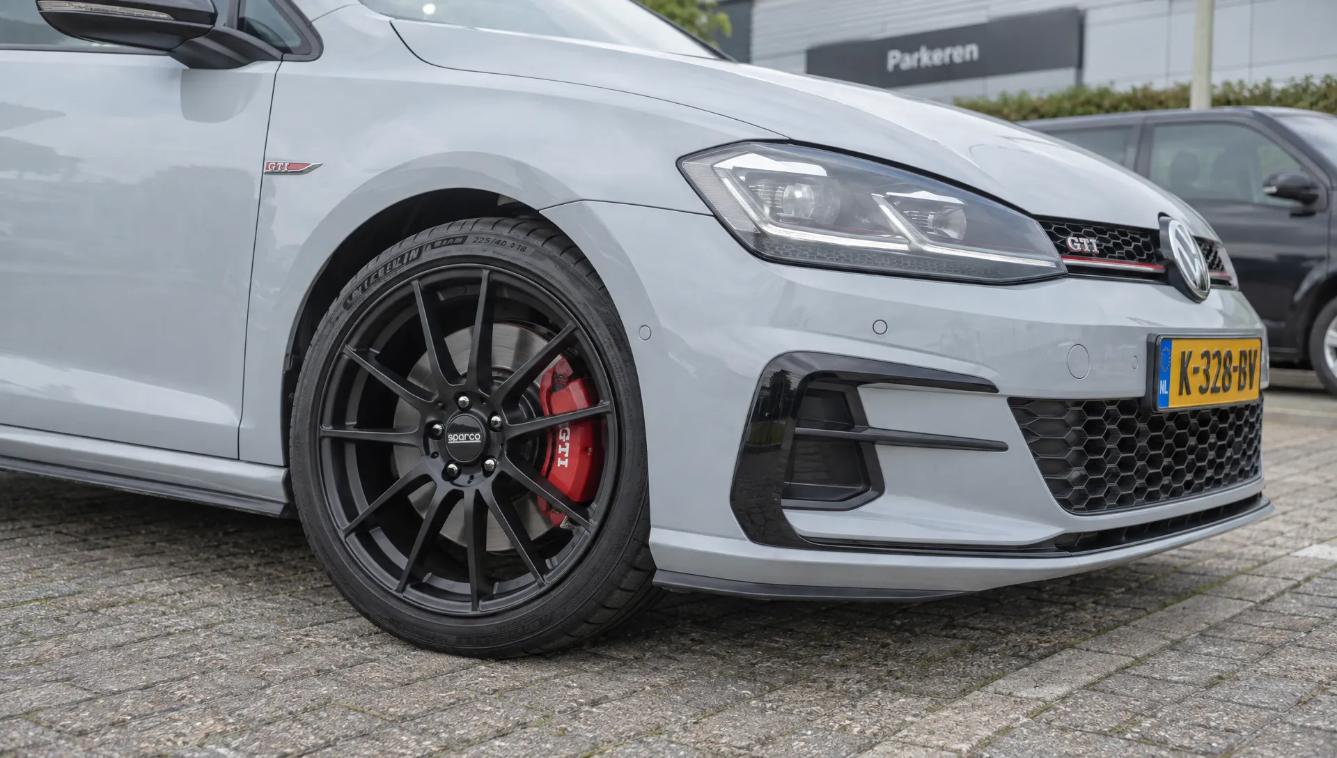 Een lichte hatchback staat op een verharde parkeerplaats met de matzwarte Sparco-velg gemonteerd; bandwang, wielkast en remklauw zijn bij het voorwiel zichtbaar.