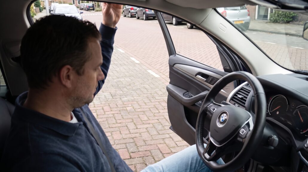 Een man zit achter het stuur van een BMW met het portier open, klaar om te rijden; de auto staat geparkeerd tussen andere auto’s.