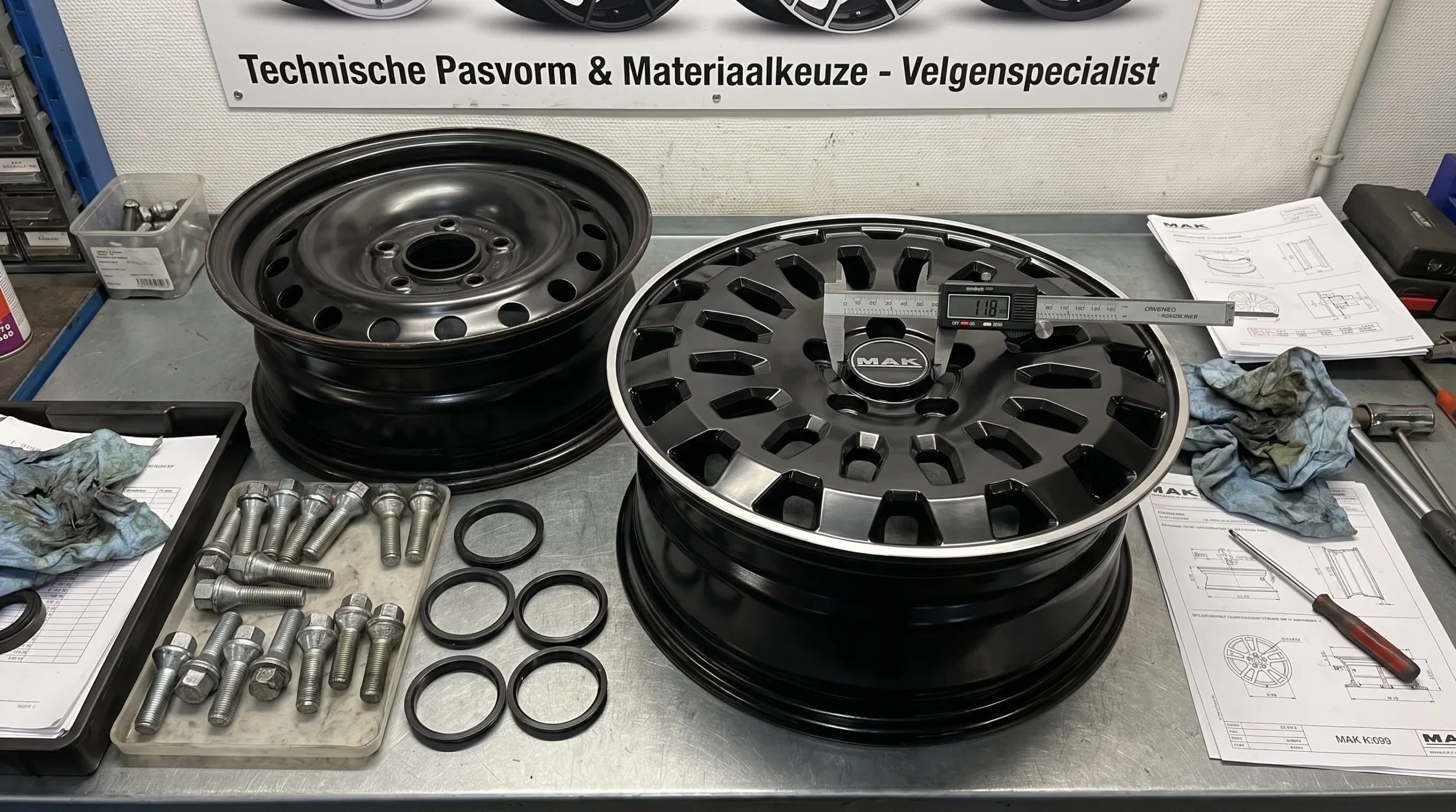 Een werkbank in een velgenspecialist-werkplaats met een lichtmetalen velg, een stalen velg, wielbouten, centreerringen en een meetinstrument, bedoeld om technische pasvorm en materiaalkeuze duidelijk te maken.