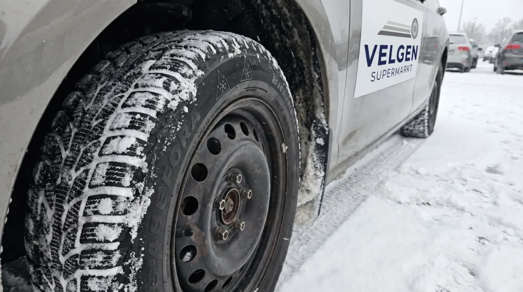 Een winterband met sneeuwvloksymbool op een stalen velg staat op een besneeuwde parkeerplaats, klaar voor de winterse rit.