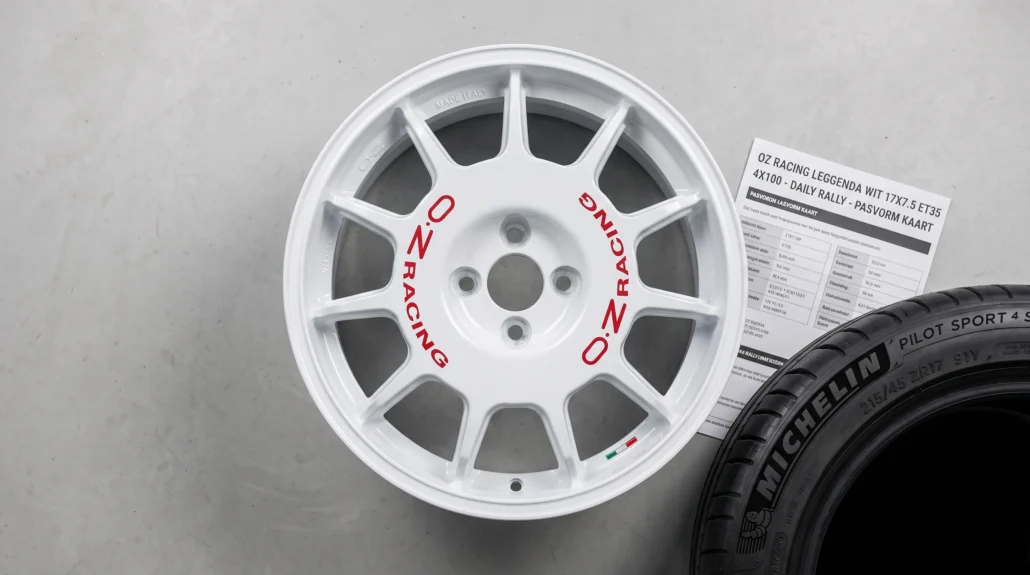 Een witte OZ rallyvelg met rode letters ligt naast een technische maatkaart en een bandenwang, zodat direct duidelijk is dat pasvorm en maat tellen.